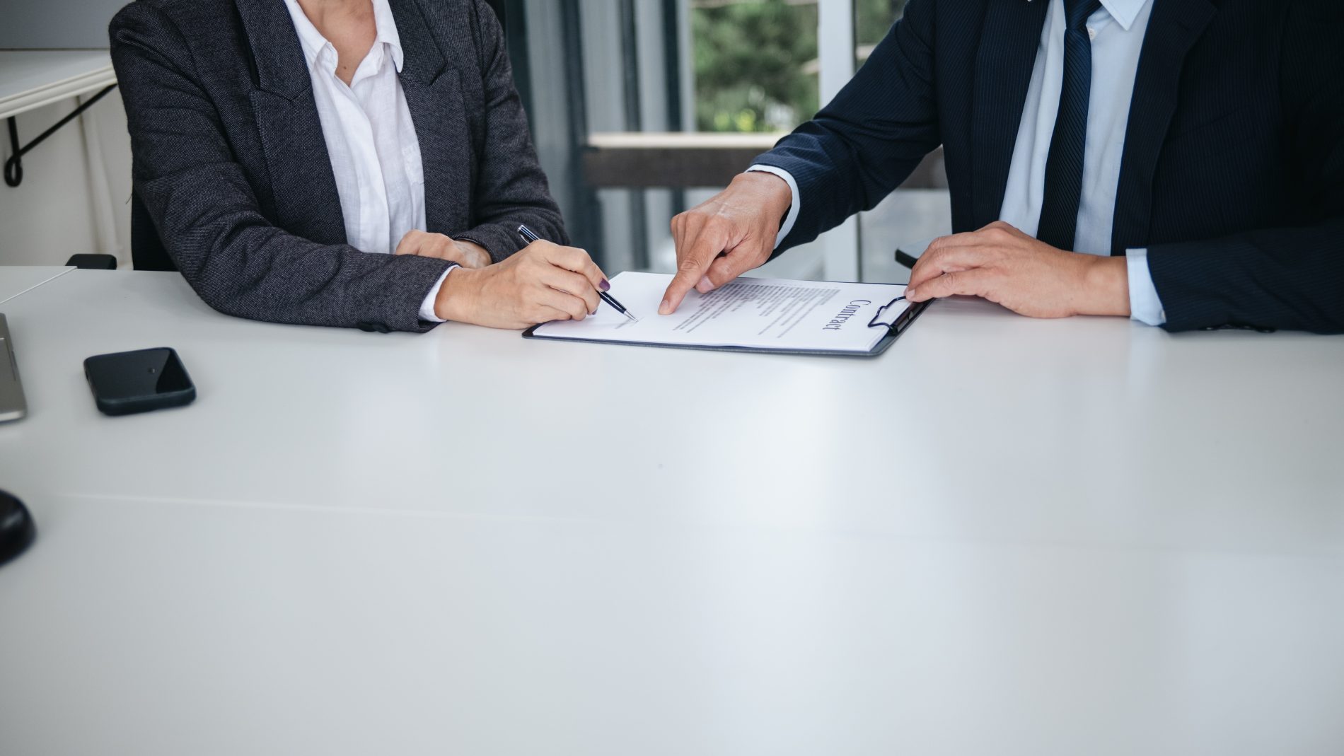 Zwei Personen im Business-Outfit sitzen an einem Tisch und besprechen ein Vertragsdokument, eine Person unterschreibt