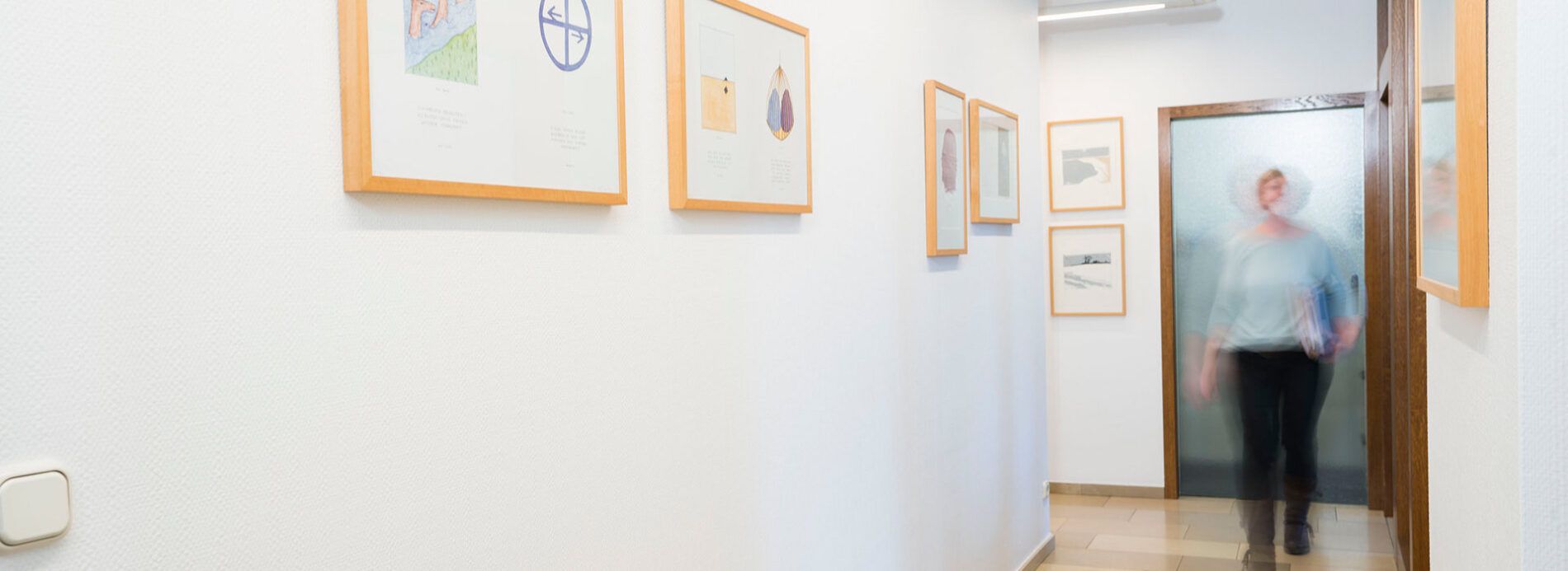 Flur im Notariat Fruh & Kerstan in Mönchengladbach mit gerahmten Bildern an der Wand und einer Person in Bewegung im Hintergrund
